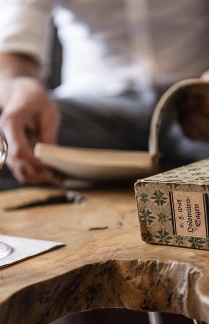 Un bicchiere con una bevanda ambrata su un tavolo in legno, accanto un libro intitolato Leggende delle Dolomiti, sullo sfondo una persona che legge.