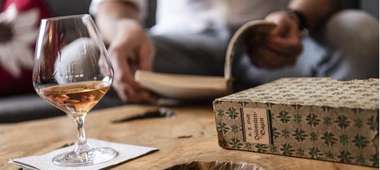A glass with an amber-colored drink on a wooden table, next to a book titled Dolomite Legends, with a person reading in the background.