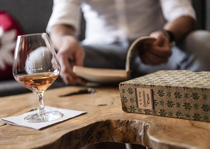 A glass with an amber-colored drink on a wooden table, next to a book titled Dolomite Legends, with a person reading in the background.