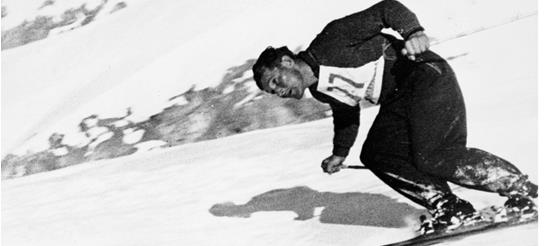 Uno sciatore con abbigliamento da sci classico del dopoguerra scende dinamicamente una pista innevata, con il pettorale del numero di partenza; foto in bianco e nero.