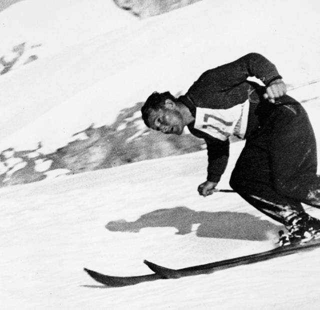 Uno sciatore con abbigliamento da sci classico del dopoguerra scende dinamicamente una pista innevata, con il pettorale del numero di partenza; foto in bianco e nero.