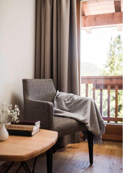 Cozy sitting area with a grey armchair, blanket, wooden table with books, and view of a balcony with wooden railing and nature in the background.
