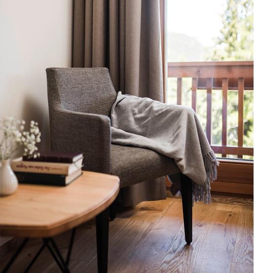 Angolo accogliente con poltrona grigia, coperta, tavolino in legno con libri e vista sul balcone con ringhiera in legno e natura sullo sfondo.