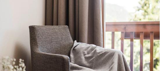 Angolo accogliente con poltrona grigia, coperta, tavolino in legno con libri e vista sul balcone con ringhiera in legno e natura sullo sfondo.