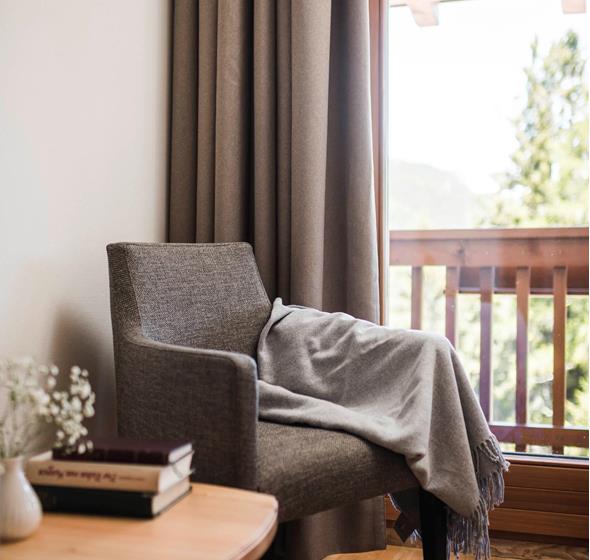 Angolo accogliente con poltrona grigia, coperta, tavolino in legno con libri e vista sul balcone con ringhiera in legno e natura sullo sfondo.