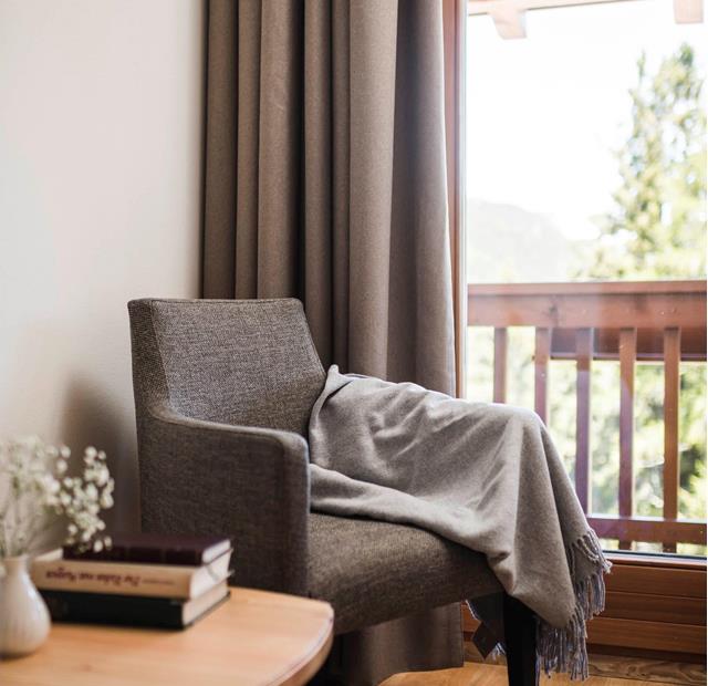 Angolo accogliente con poltrona grigia, coperta, tavolino in legno con libri e vista sul balcone con ringhiera in legno e natura sullo sfondo.
