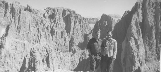 Due uomini in abiti da escursionismo tradizionali posano su una sporgenza rocciosa con una vista spettacolare sulle frastagliate cime delle Dolomiti sullo sfondo; foto in bianco e nero.