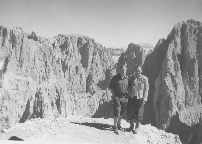 Zwei Männer in traditioneller Wanderkleidung posieren auf einem Felsvorsprung mit dramatischer Aussicht auf die zerklüfteten Gipfel der Dolomiten im Hintergrund; Schwarzweißaufnahme.