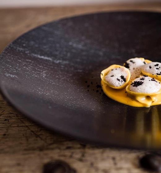 Three filled tortellini served on a creamy orange sauce, topped with foam and black crumbs, presented on a rustic dark plate.