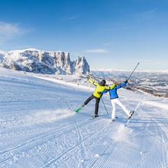 Zwei Skifahrer in bunter Winterkleidung feiern ausgelassen auf einer frisch präparierten Piste mit Blick auf die verschneiten Dolomiten und das weite Tal im Hintergrund.