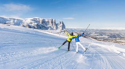 Zwei Skifahrer in bunter Winterkleidung feiern ausgelassen auf einer frisch präparierten Piste mit Blick auf die verschneiten Dolomiten und das weite Tal im Hintergrund.
