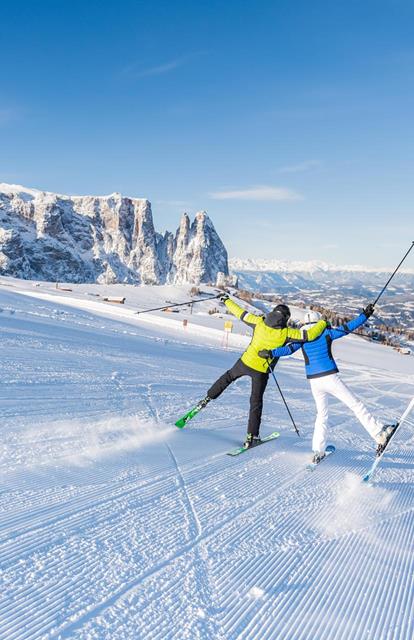 Due sciatori in abbigliamento invernale colorato festeggiano su una pista appena battuta con vista sulle Dolomiti innevate e la valle sullo sfondo.