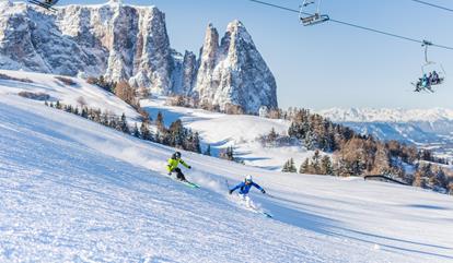 Zwei Skifahrer auf breiter Piste, Sessellifte darüber, mit schneebedeckten Dolomiten im Hintergrund.