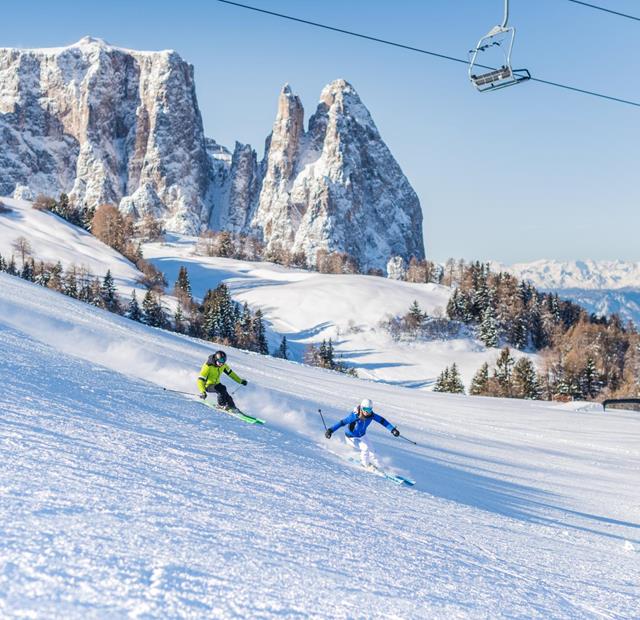 Due sciatori su una pista larga, seggiovie sopra, con le Dolomiti innevate sullo sfondo.