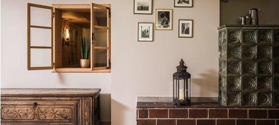 Traditionell eingerichteter Raum mit grüner Kachelheizung, antiker Holztruhe, offener Innenfensteröffnung mit Pflanze und historischen Schwarz-Weiß-Fotografien an der Wand.