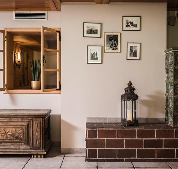 Traditionally furnished room with green tiled stove, antique wooden chest, open interior window with a plant, and historical black-and-white photographs on the wall.