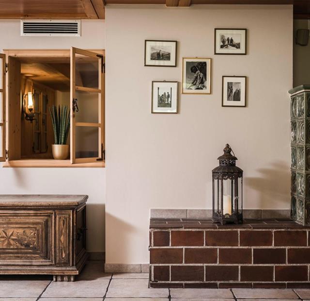 Traditionell eingerichteter Raum mit grüner Kachelheizung, antiker Holztruhe, offener Innenfensteröffnung mit Pflanze und historischen Schwarz-Weiß-Fotografien an der Wand.